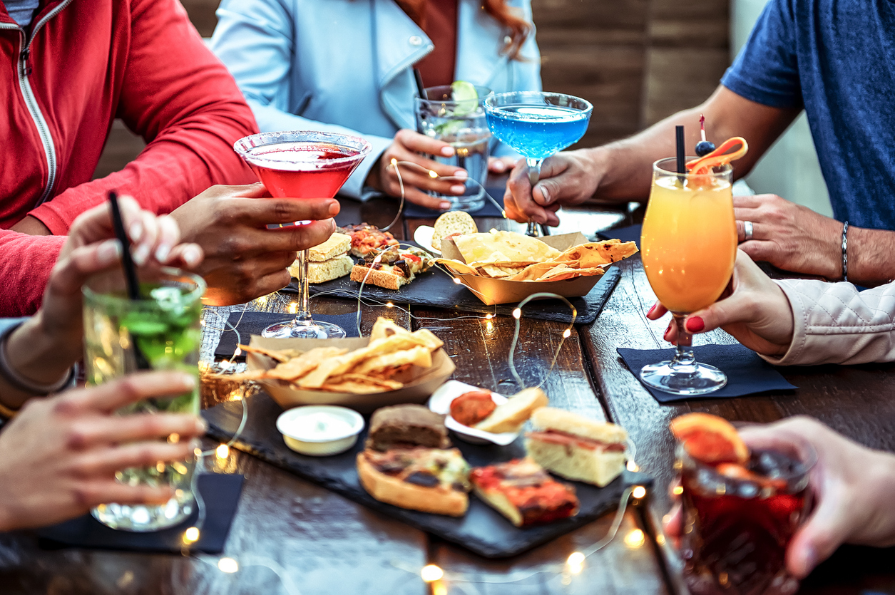 Group of friends enjoying appetizer drinking and eating in a bar - Close-up of hands of young people holding colorful cocktails in the happy hour time - Social gathering party time concept